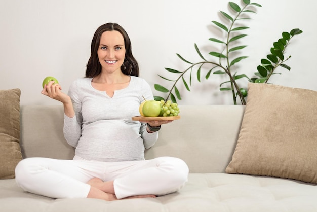 Donna incinta sorridente seduta sul divano con piatto di frutta sul braccio sano concetto di gravidanza
