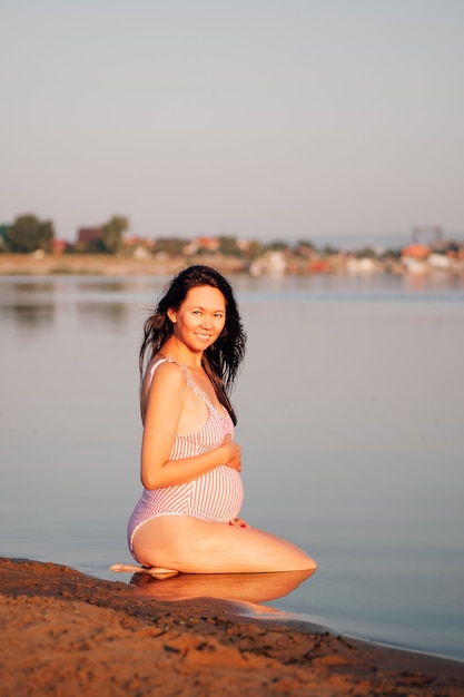 Donna incinta sulla spiaggia pacifica donna incinta asiatica in un costume da bagno rosa è seduta sulle sue ginocchia...