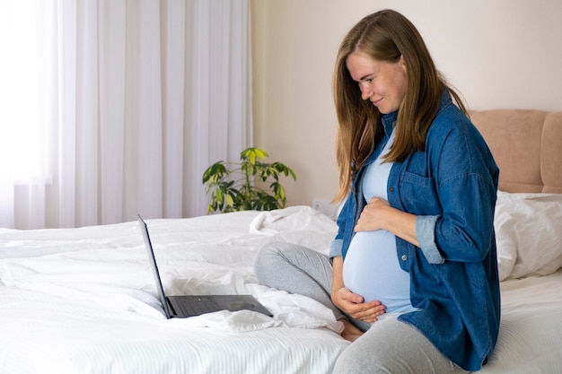Donna incinta usa il suo laptop a letto a casa navigando in rete fare shopping online leggendo blog di maternità o chattando online Gravidanza e tecnologia