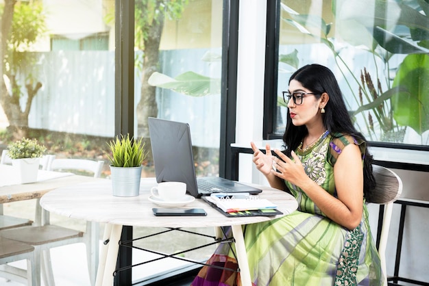 Donna indiana con occhiali utilizzando un computer portatile con un notebook e un telefono cellulare con una tazza di caffè sul tavolo