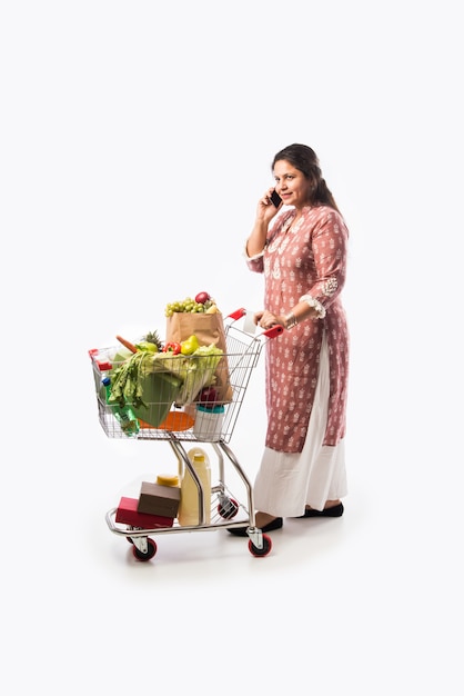 Donna indiana di mezza età con carrello della spesa o carrello pieno di generi alimentari, frutta e verdura. Isolato Foto a figura intera su muro bianco