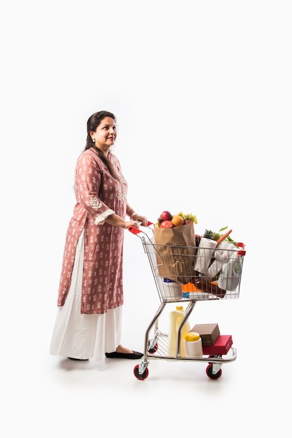 Donna indiana di mezza età con carrello della spesa o carrello pieno di generi alimentari, frutta e verdura. Isolato Foto a figura intera su muro bianco