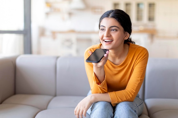 Donna indiana sorridente che registra un messaggio vocale sullo smartphone mentre si rilassa a casa