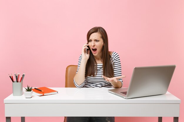 Donna indignata che allarga le mani parlando al cellulare mentre è seduta, lavorando su un progetto in ufficio con un computer portatile isolato su sfondo rosa pastello. Concetto di carriera aziendale di successo. Copia spazio.