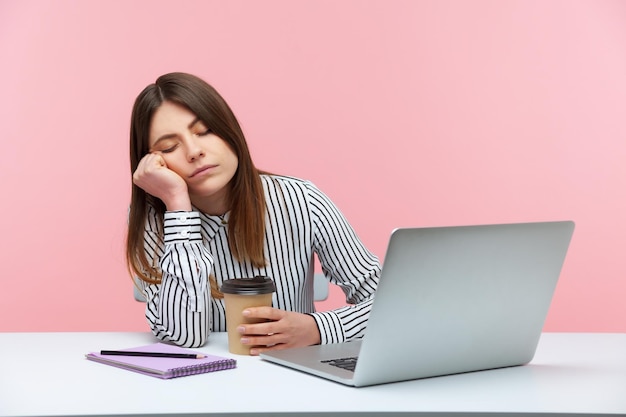 Donna inefficiente assonnata che fa un pisolino con la testa appoggiata a portata di mano e tiene una tazza di caffè seduta sul posto di lavoro con esaurimento fisico del laptop Colpo in studio al coperto isolato su sfondo rosa