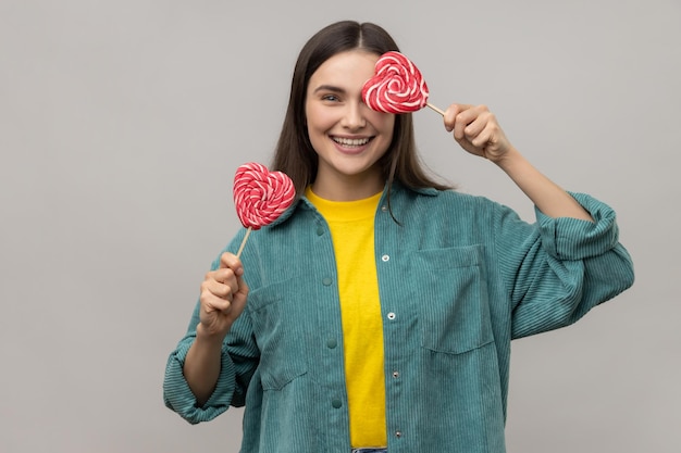 Donna infantile con i capelli scuri che copre l'occhio con lecca-lecca a forma di cuore che si nasconde divertendosi con i dolci