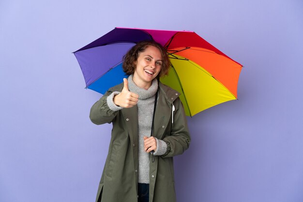 Donna inglese che tiene un ombrello isolato su sfondo viola con i pollici in su perché è successo qualcosa di buono