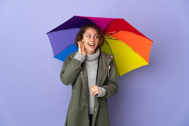Donna inglese che tiene un ombrello isolato sulla parete viola ascoltando qualcosa mettendo la mano sull'orecchio