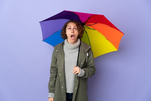 Donna inglese che tiene un ombrello isolato sulla parete viola che osserva in su e con l'espressione sorpresa