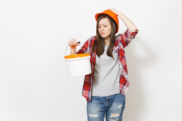 Donna insoddisfatta triste in elmetto protettivo che guarda sul secchio vuoto della vernice con lo spazio della copia e aggrappandosi alla testa isolata su fondo bianco. Strumento per ristrutturazione appartamento. Riparare il concetto di casa.