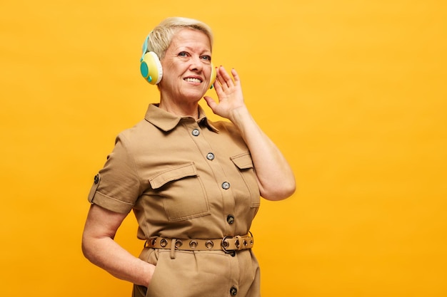 Donna invecchiata felice in vestito beige alla moda che ascolta la musica in cuffie
