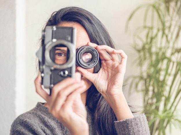 Donna irriconoscibile con lunghi capelli scuri che guarda attraverso il foro nella fotocamera analogica retrò mentre copre l'occhio con la lente e finge di scattare una foto