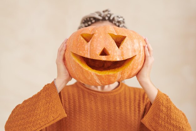 Donna irriconoscibile in maglione marrone che tiene una zucca divertente contro la festa di Halloween del viso