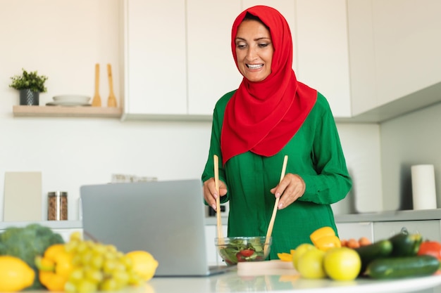 Donna islamica matura che prepara insalata e guarda il computer portatile in piedi nella cucina moderna di casa