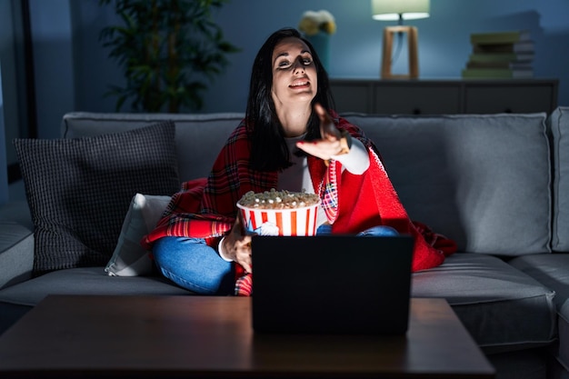 Donna ispanica che mangia popcorn guardando un film sul divano sorridendo amichevole offrendo la stretta di mano come saluto e benvenuto. business di successo.