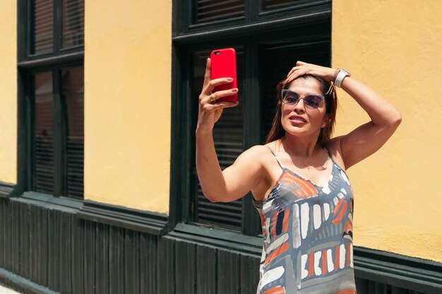 Donna ispanica con capelli lunghi e occhiali che cammina per le strade della città e si fa un selfie con il cellulare.