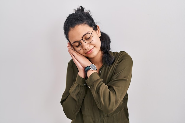 Donna ispanica con capelli scuri in piedi su sfondo isolato dormire stanco sognare e posare con le mani insieme mentre sorride con gli occhi chiusi