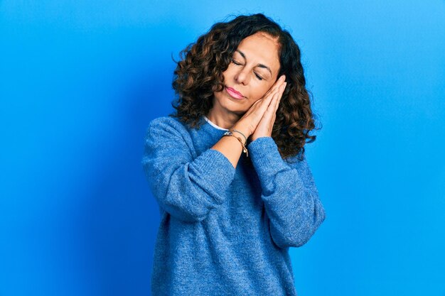 Donna ispanica di mezza età che indossa abiti casuali che dorme stanca che sogna e posa con le mani unite mentre sorride con gli occhi chiusi