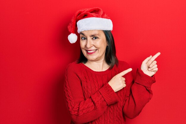 Donna ispanica di mezza età che indossa il cappello di Natale sorridente e guardando la telecamera che punta con due mani e le dita di lato