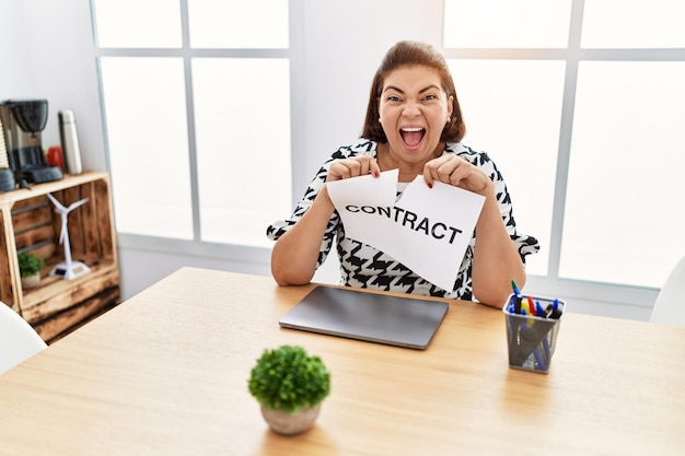 Donna ispanica di mezza età che rompe la carta del contratto in ufficio festeggiando pazza e stupita per il successo con gli occhi aperti urlando eccitata