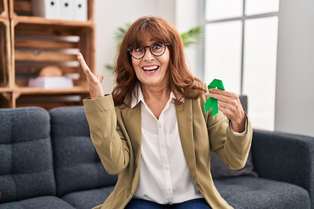 Donna ispanica di mezza età che tiene il nastro verde di supporto che celebra la vittoria con un sorriso felice e l'espressione del vincitore con le mani alzate