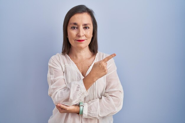Donna ispanica di mezza età in piedi su sfondo blu che punta con il dito della mano verso il lato che mostra pubblicità, viso serio e calmo