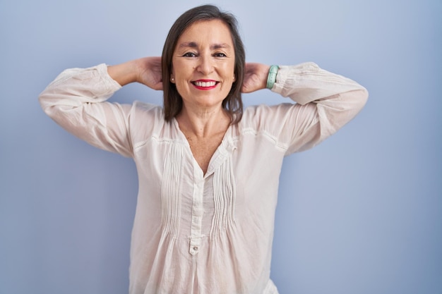 Donna ispanica di mezza età in piedi su sfondo blu rilassante e allungando le braccia e le mani dietro la testa e il collo sorridendo felice