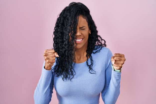 Donna ispanica di mezza età in piedi su sfondo rosa entusiasta per il successo con le braccia alzate e gli occhi chiusi che celebrano la vittoria sorridendo. concetto di vincitore.