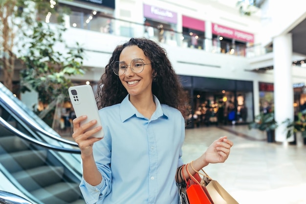 Donna ispanica felice nello shopping del supermercato che legge il messaggio online che tiene le borse della spesa colorate