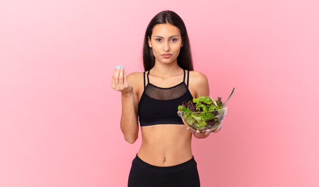 Donna ispanica fitness che fa un gesto capice o denaro, dicendoti di pagare e tenendo in mano un'insalata