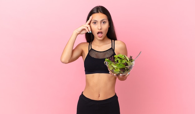 Donna ispanica fitness che sembra sorpresa, realizzando un nuovo pensiero, idea o concetto e tenendo in mano un'insalata