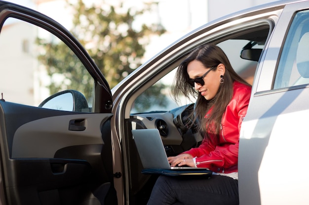 Donna ispanica in occhiali da sole che lavora con un computer portatile dalla sua auto