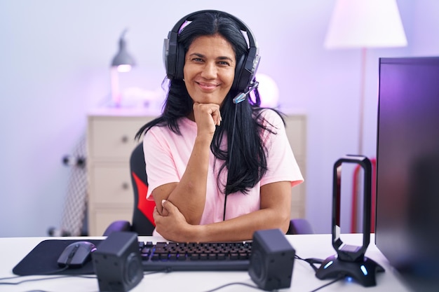 Donna ispanica matura che gioca ai videogiochi a casa guardando fiduciosa alla telecamera sorridendo con le braccia incrociate e la mano alzata sul mento pensando positivo