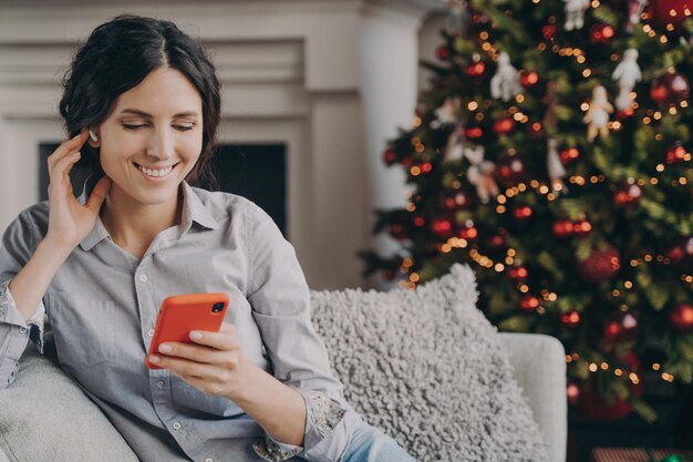 Donna italiana rilassata e felice seduta su un comodo divano a casa con il telefono durante le vacanze di natale