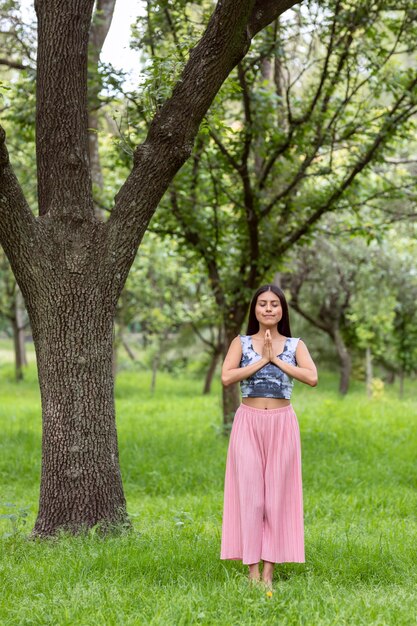 Donna latina che fa meditazione sul lato di un albero nel parco su erba verde con un vestito rosa