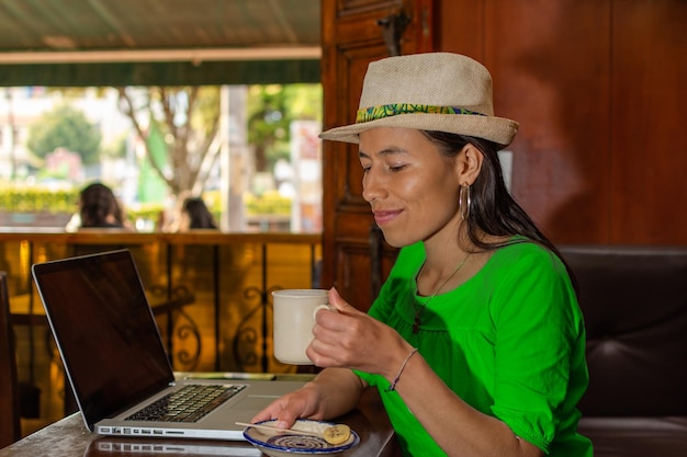 Donna latina che indossa un cappello seduto in una vecchia caffetteria che lavora al suo computer portatile con in mano una tazza di caffè