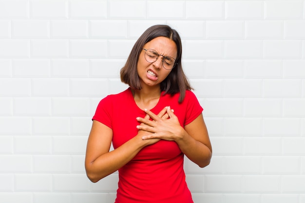 Donna latina che sembra triste, ferita e affranta, tenendo entrambe le mani vicino al cuore, piangendo e sentendosi depressa