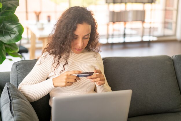 Donna latina che utilizza laptop e mano che tiene la carta di credito per lo shopping sul divano di casa