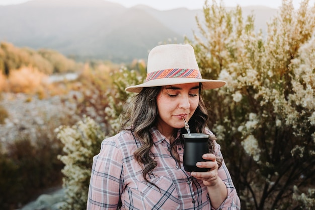 Donna latina con un cappello e indossa una camicetta rosa pallido, bevendo compagno in uno spazio naturale al tramonto