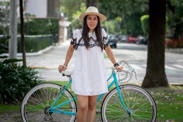 Donna latina con una bicicletta che indossa vestito e cappello in un parco