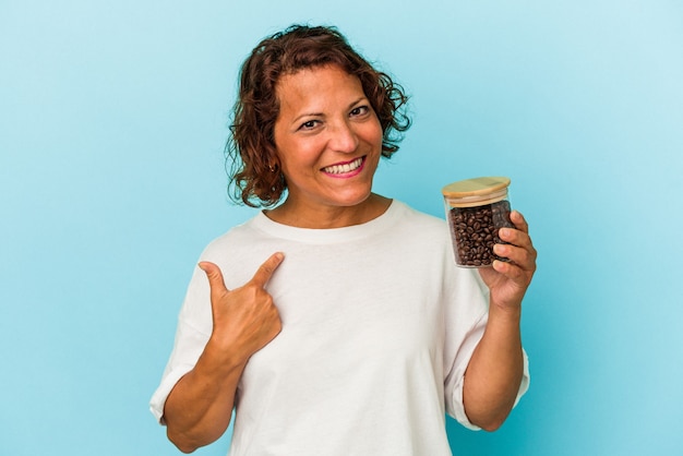 Donna latina di mezza età che tiene in mano un barattolo di caffè isolato su sfondo blu che punta con il dito verso di te come se invitasse ad avvicinarsi.