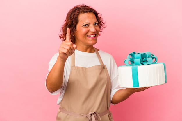 Donna latina di pasticceria di mezza età che tiene una torta isolata su sfondo rosa che mostra il numero uno con il dito.