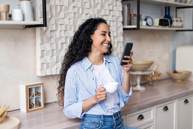 Donna latina eccitata che usa lo smartphone mentre beve il tè del mattino in cucina a casa leggendo i messaggi