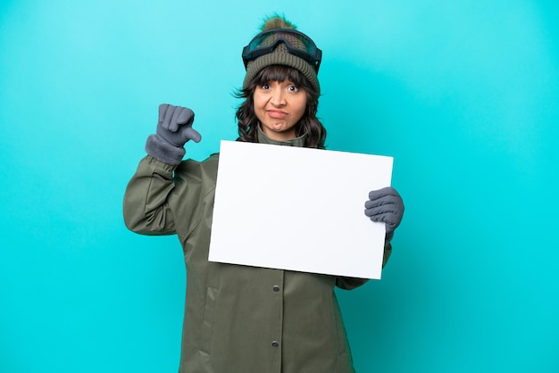 Donna latina sciatore con occhiali da snowboard isolati su sfondo blu che tiene un cartello vuoto e fa un cattivo segnale