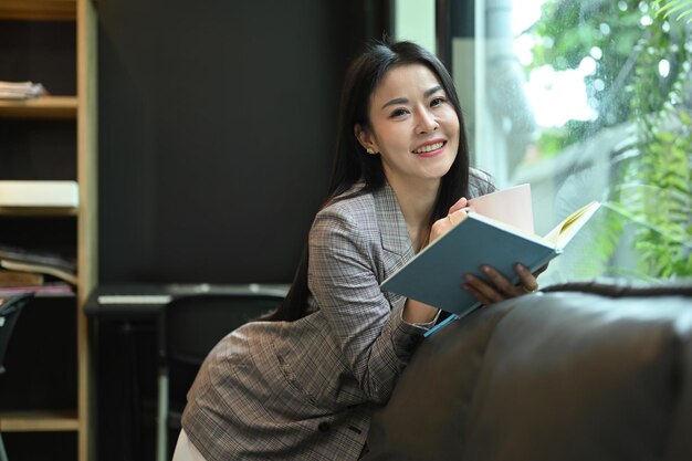 Donna lavoratrice asiatica allegra che si rilassa sul divano nel suo ufficio moderno luminoso e libro di lettura