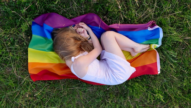 Donna lesbica sdraiata sulla bandiera LGBT sull'erba in abito bianco Supporto dell'orientamento non tradizionale nel mese della dignità il giorno della bisessualità La persona manca e nasconde il viso dal bullismo