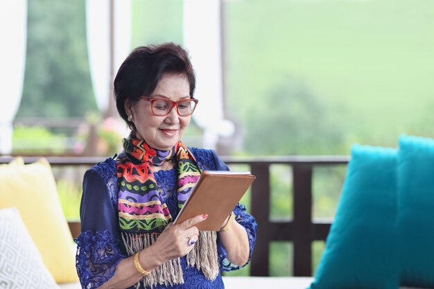 Donna maggiore asiatica che si siede sul divano e utilizzando un tablet