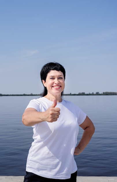 Donna maggiore attiva e felice in abiti sportivi che mostra i pollici in su che lavora vicino alla riva del fiume