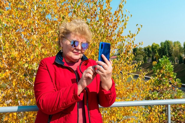 Donna maggiore caucasica in giacca rossa utilizza lo smartphone nel parco