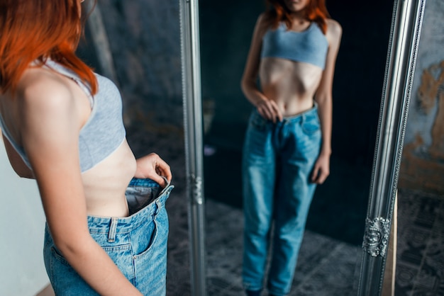 Donna magra prova jeans di grandi dimensioni contro specchio, perdita di peso, anoressia. Concetto di bruciare calorie o grassi, malattia medica